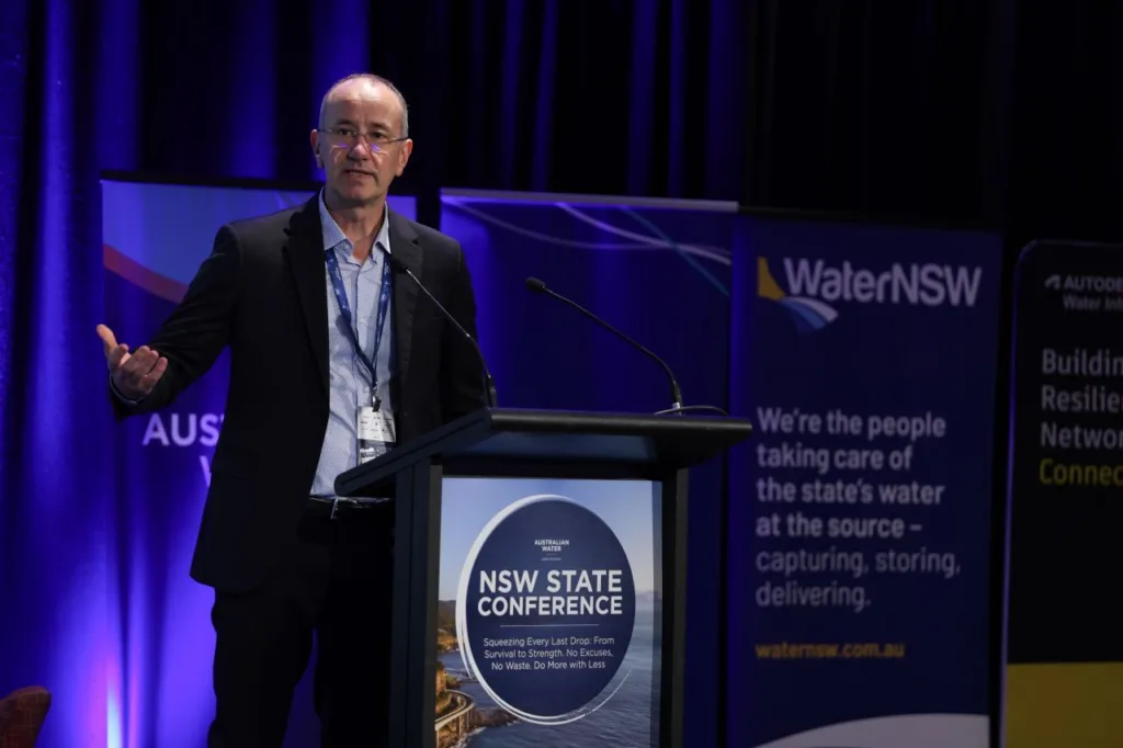 A man in a suit speaks at the lectern during the WaterNSW State Conference, gesturing towards the audience.
